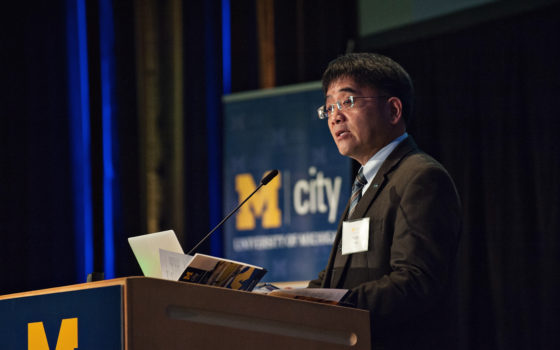 Professor Huei Peng stands at podium with Mcity banner behind his right shoulder. Banner is blue with white print. Professor Peng has black hair and wears glasses. He is wearing brown jacket, white shirt and tie.