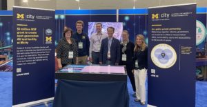 U-M team members standing in exhibit at AutoMobili-D 2022, flanked by two blue and gold banners about Mcity.