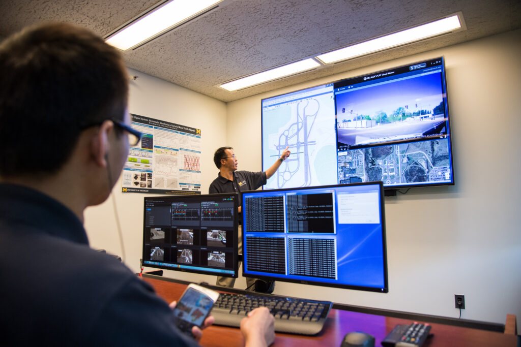 Henry Liu, wearing a blue polo shirt with Mcity logo on right chest, points to large display monitor showing a schematic and images of the Mcity Test Facility