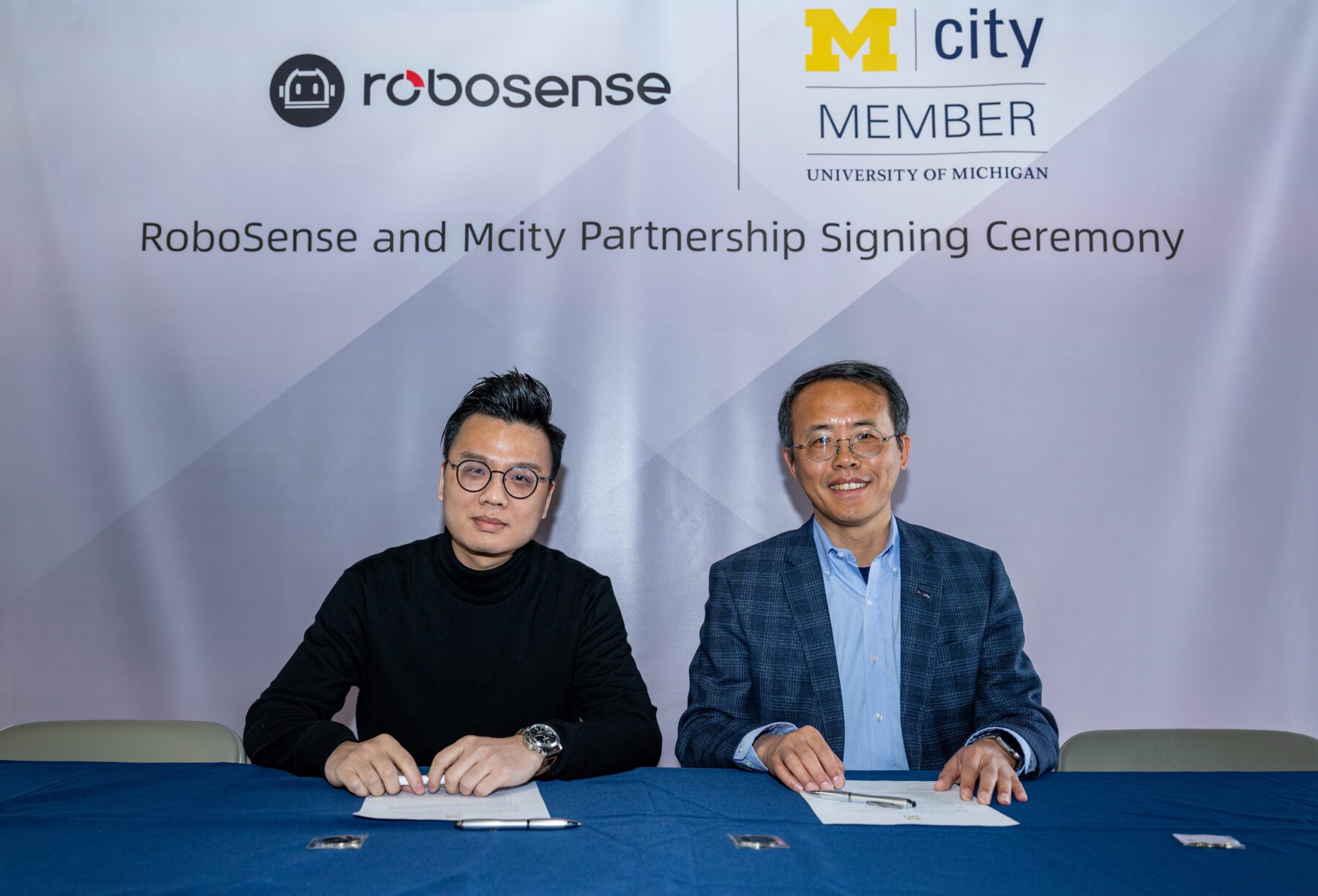 Two Asian men sit at a table with white papers and pens in front of them. Behind them is a banner that says, "RoboSense and Mcity Partnership Signing Ceremony."
