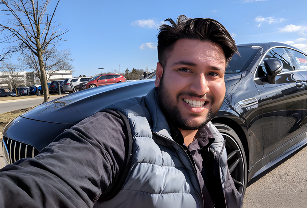 A young man with dark hair and brown skin from India takes a selfie in front of a vehicle.