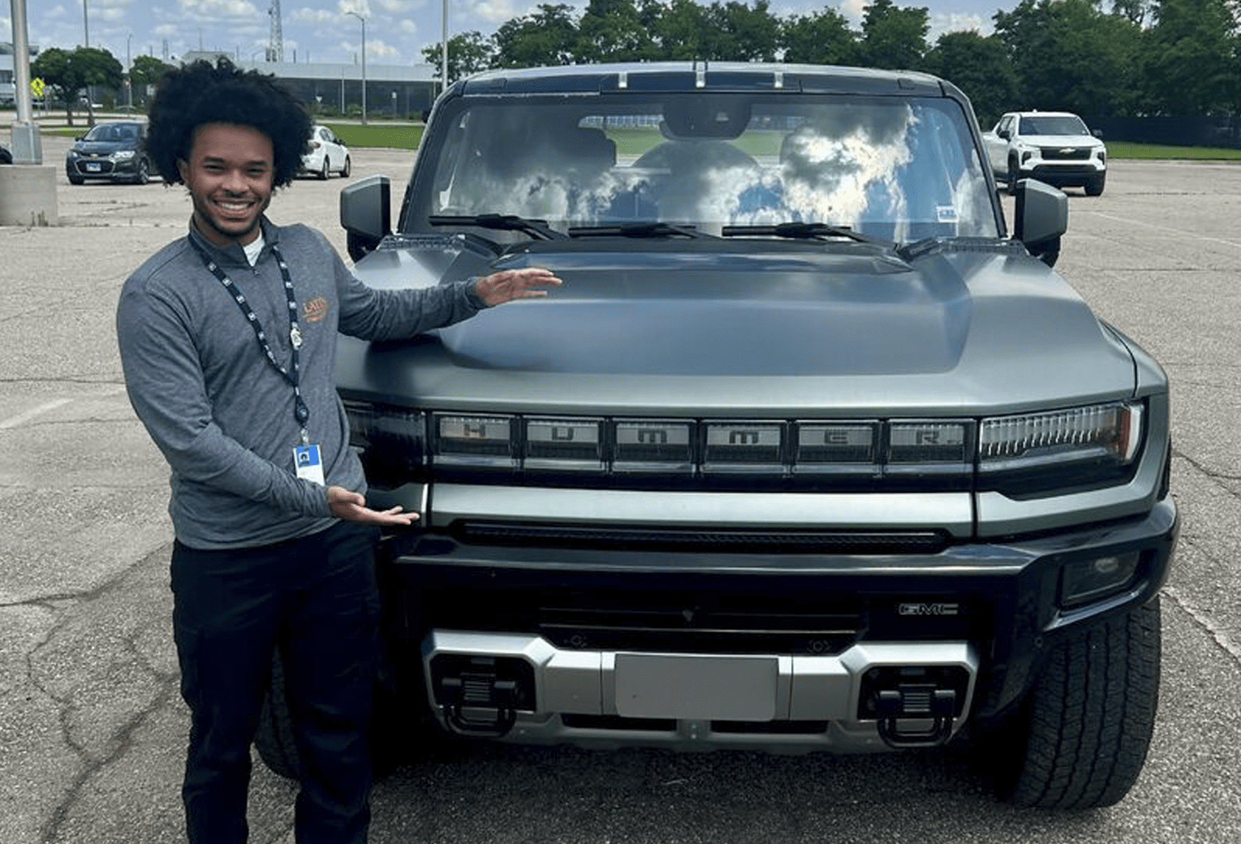 A young black man in dark pants and a gray shirt stands in front of a Hummer SUV, made by General Motors, with his arms open.