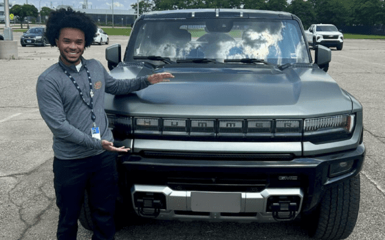 A young black man in dark pants and a gray shirt stands in front of a Hummer SUV, made by General Motors, with his arms open.