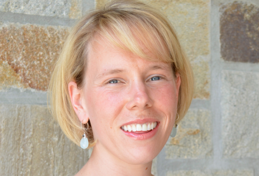 Woman with mid-length blond hair and blue eyes smiles while standing in front of a brown and tan brick wall