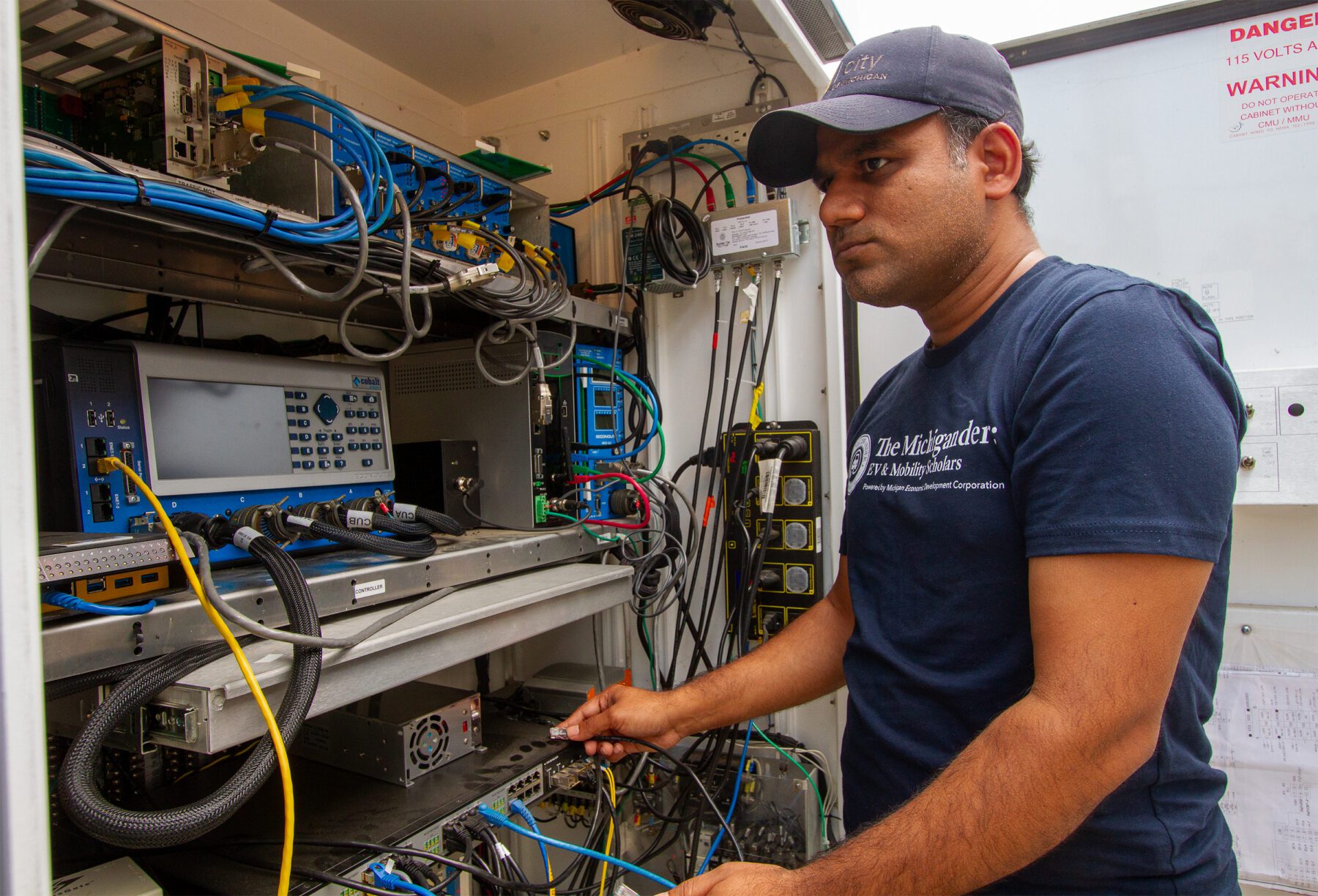 Man wearing a dark blue shirt with white lettering that says, "The Michigander EV & Mobility Scholars" stands in front of a cabinet that stores traffic control equipment at the Mcity Test Facility.