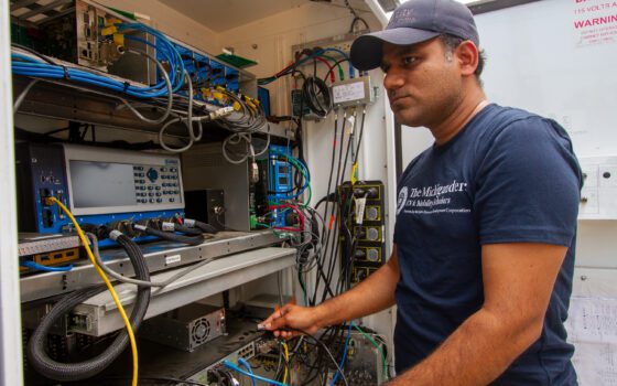 Man wearing a dark blue shirt with white lettering that says, "The Michigander EV & Mobility Scholars" stands in front of a cabinet that stores traffic control equipment at the Mcity Test Facility.