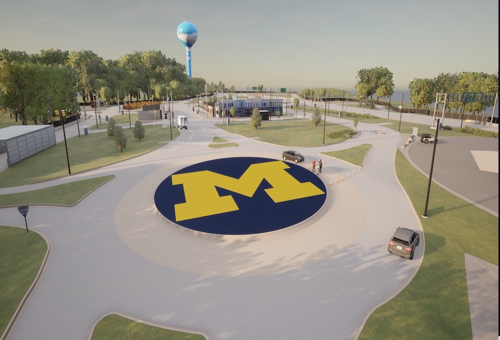 The simulation shows Mcity's roundabout with the block M in the center and green grass surrounding. The roundabout has multiple access roads and is partially blocked by a set of cones with some pedestrians nearby. More test track extends into the distance.