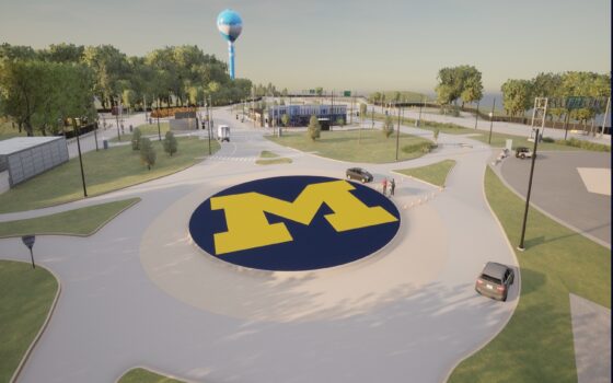 The simulation shows Mcity's roundabout with the block M in the center and green grass surrounding. The roundabout has multiple access roads and is partially blocked by a set of cones with some pedestrians nearby. More test track extends into the distance.