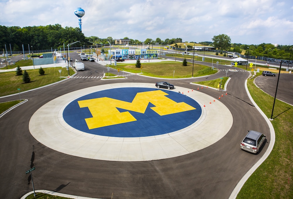 Mcity's roundabout with the block M in the center and green grass surrounding. The roundabout has multiple access roads and is partially blocked by a set of cones with some pedestrians nearby. More test track extends into the distance.