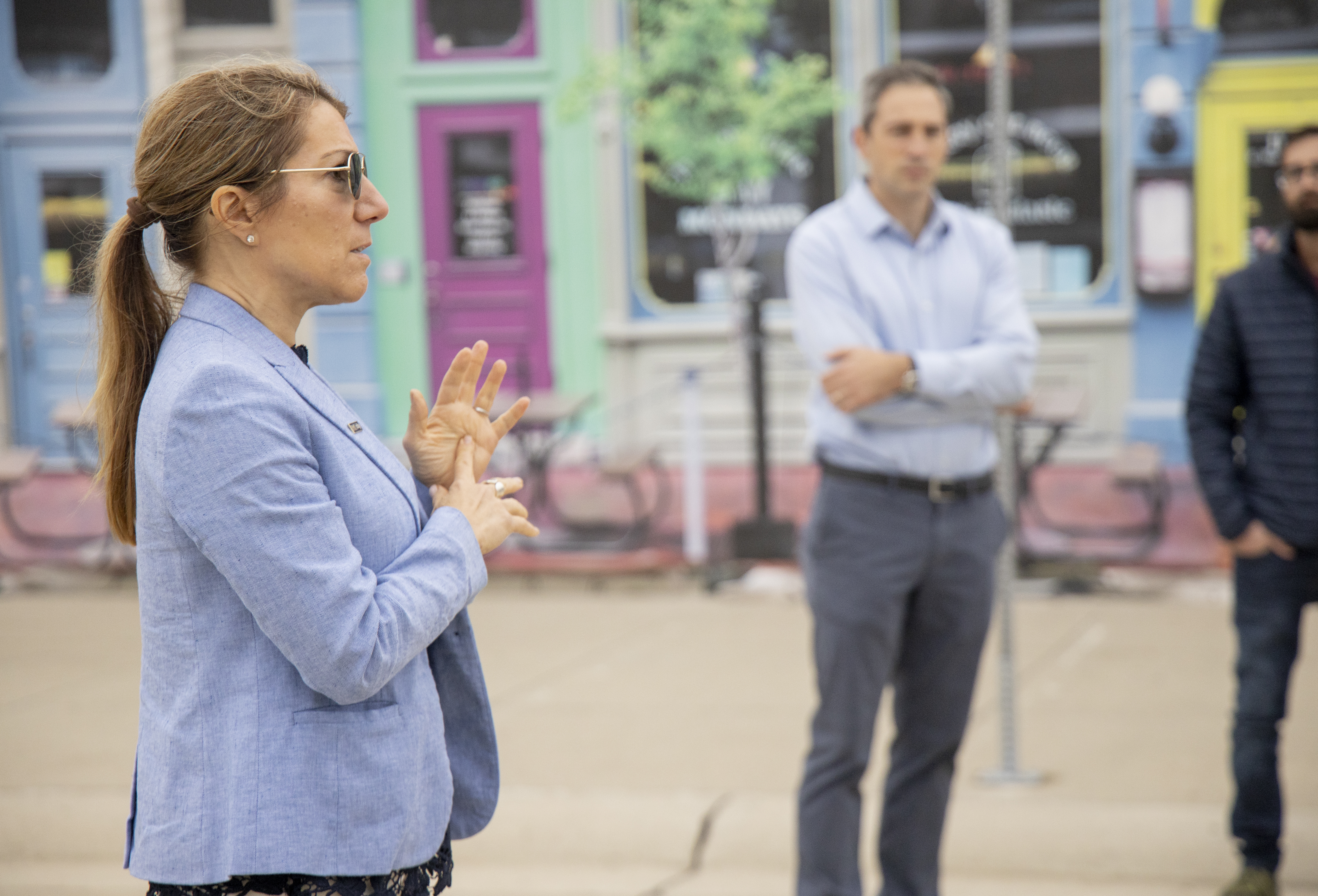 A woman with dark blond hair pulled back in a ponytail and wearing sunglasses speaks to a group of people during a tour of the Mcity Test Facility. A facade showing a downtown scene of Ann Arbor, MI is in the background.