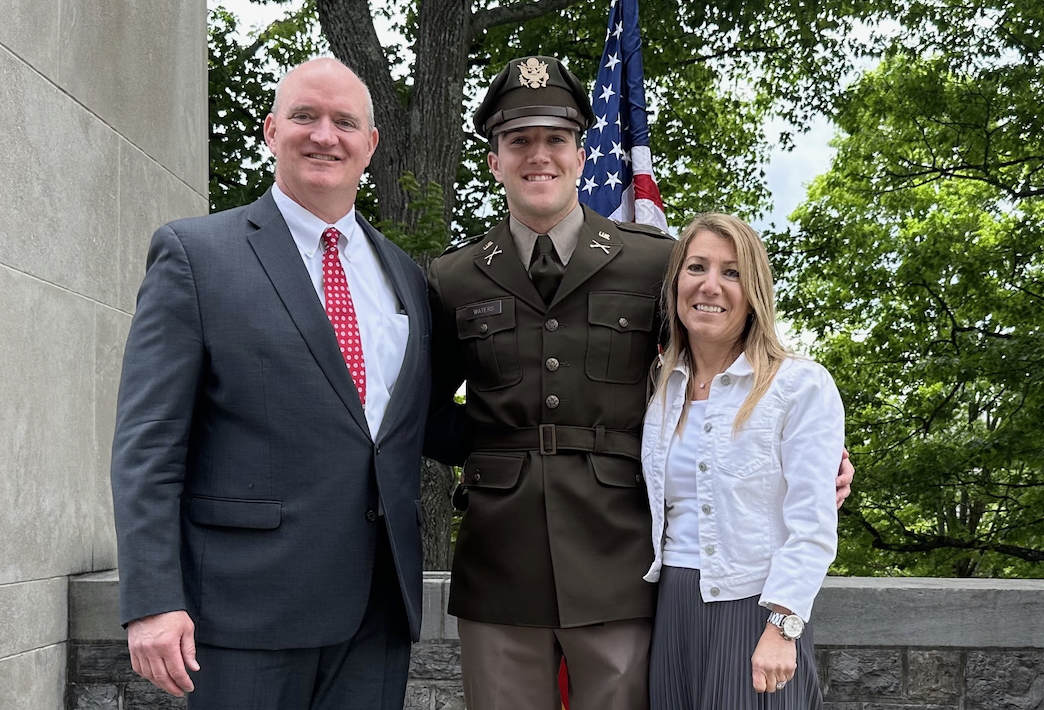 Tall man in blue suit, light shirt and red tie stands next to younger man in green Army uniform, who is standing next to a woman in a dark skirt and white shirt and denim jacket.