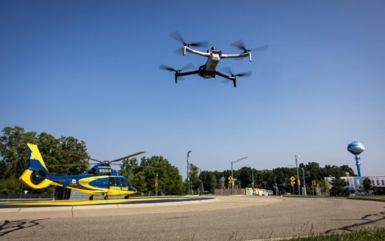 Medical helicopter that says Survival Flight in dark gold letters against dark blue background is on the ground at the Mcity Test Facility while a gray drone hovers overhead.