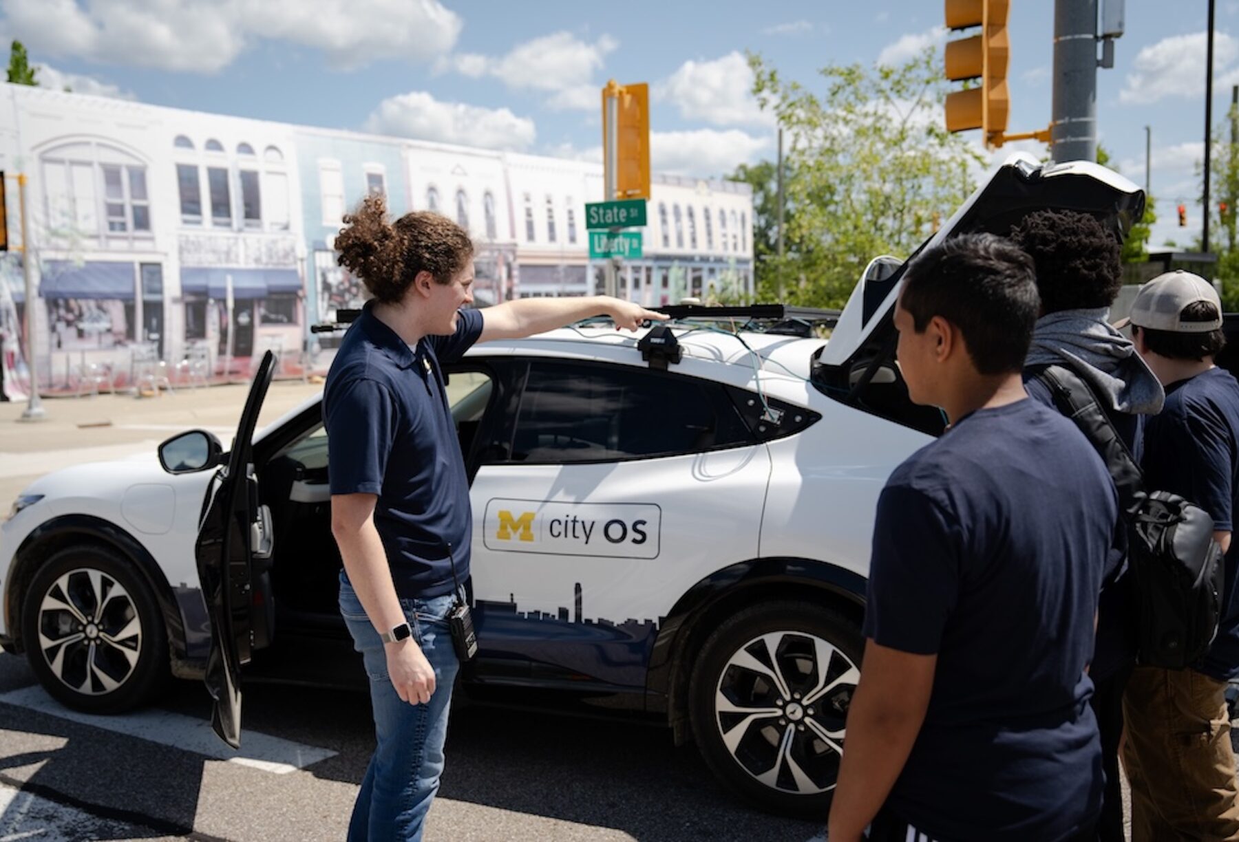 A man with dark curly hair in a pony tail points out features on a Ford Mach-E research vehicle to middle school students visiting the Mcity Test Facility to learn more about autonomous vehicles and the future of mobility.
