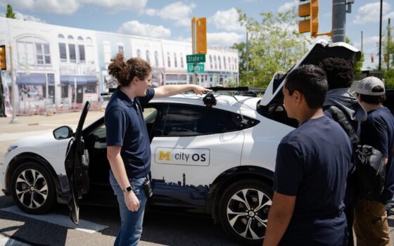 A man with dark curly hair in a pony tail points out features on a Ford Mach-E research vehicle to middle school students visiting the Mcity Test Facility to learn more about autonomous vehicles and the future of mobility.