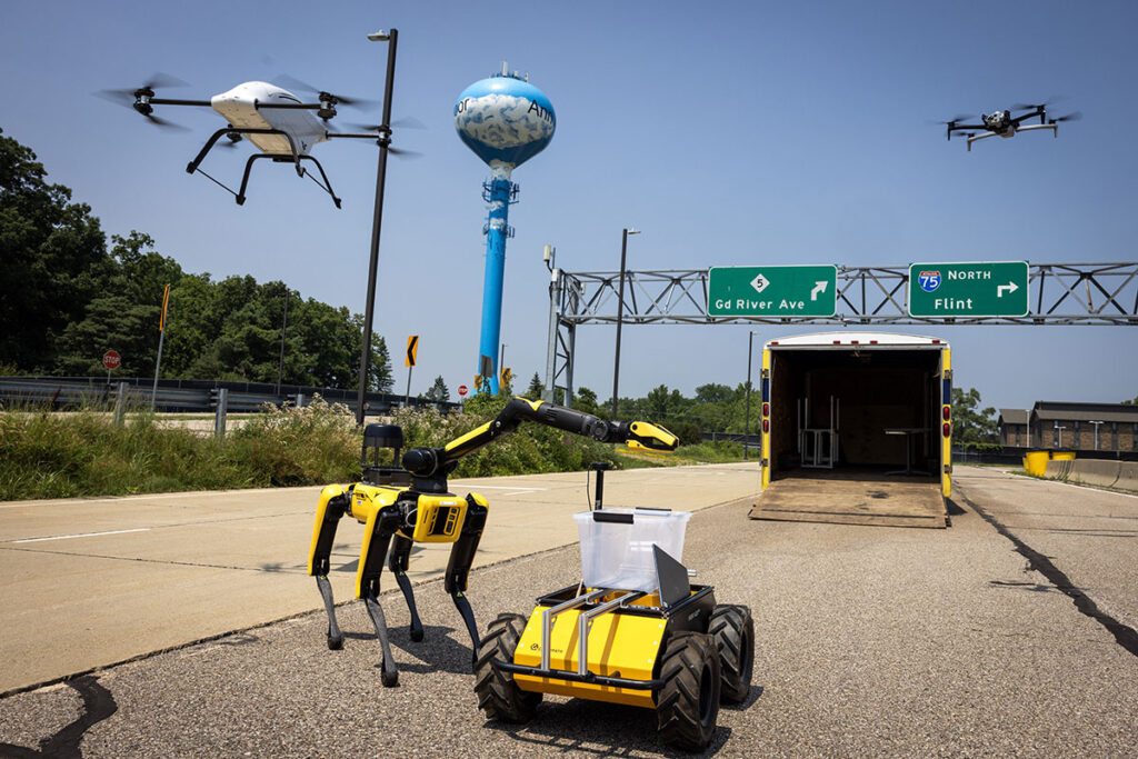 A pair of drones—including a white Cobalt cargo delivery drone, used by Brighton, Michigan-based company blueflite—and two ground robots operated by University of Michigan staff conduct tests at the Mcity Test Facility on U-M's North Campus.